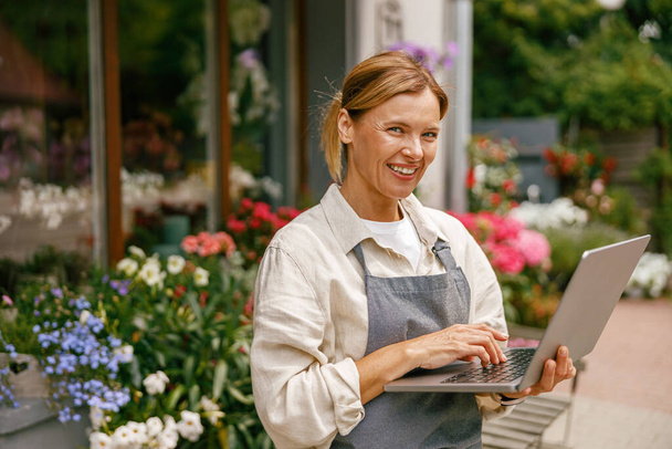 Bonita dona de loja de flores vestindo avental trabalhando em laptop em sua loja - Foto, Imagem