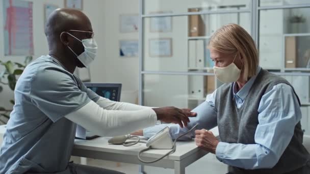 Młody Afroamerykański lekarz w mundurze i masce ochronnej z użyciem tonometru, siedzący przy pracy w szpitalu i mierzący ciśnienie krwi dojrzałej pacjentki podczas konsultacji - Materiał filmowy, wideo