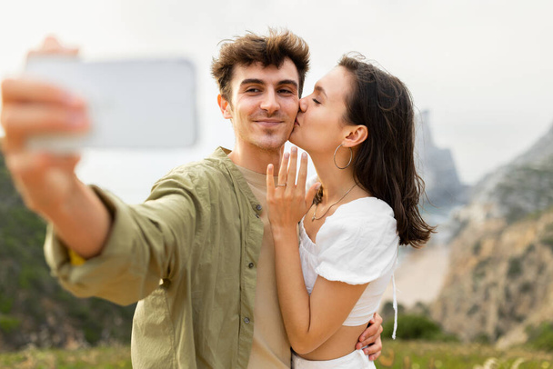 Happy loving couple after engagement taking selfie on cellphone, lady kissing man in cheek, capturing perfect beachfront proposal, standing on rocks cliff near ocean shore - Photo, Image
