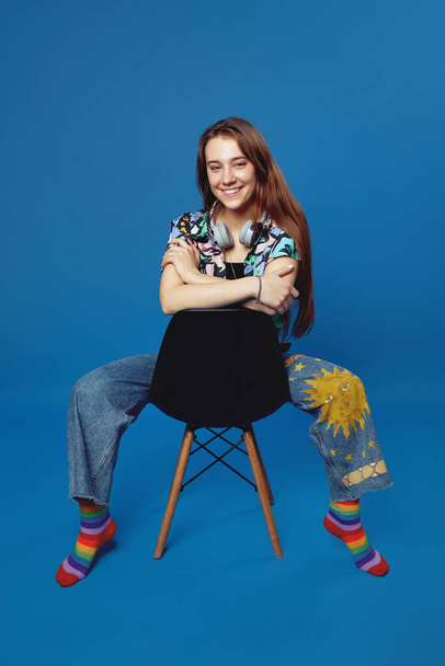 Vertical photo of excited student girl with headphones wearing trendy shirt and jeans, looking at camera while sitting on chair against blue background. - Φωτογραφία, εικόνα