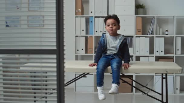 Schattige kleine jongen in spijkerjasje en blauwe spijkerbroek zittend op de bank in het centrum van het medisch kantoor van moderne klinieken en kijkend naar de camera terwijl zwaaiende benen en grimmig - Video