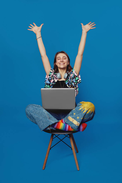 Χαρούμενη μαθητριούλα με ακουστικά στο λαιμό κάθεται στην καρέκλα με το laptop, σηκώνοντας τα χέρια και κάνει χειρονομία νίκης, απομονωμένη σε μπλε φόντο. - Φωτογραφία, εικόνα