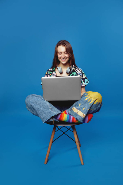 Photo verticale de fille étudiante heureuse jouie en chemise colorée, bavarder avec des amis par ordinateur portable, assis avec les jambes croisées sur la chaise tout en posant isolé sur fond de studio bleu. - Photo, image