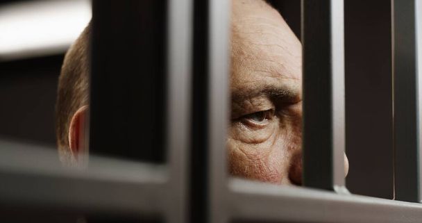 Hapishane hücresinde metal parmaklıkların arkasında duran depresif yaşlı mahkumun yüzüne yakından bakın. Suçlu ya da katil, suçtan hapis yatıyor. Hapiste ya da gözaltı merkezinde. Adalet sistemi. - Fotoğraf, Görsel