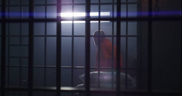 Літній злочинець у помаранчевій формі сидить у в'язниці, встає і дивиться на вікно з брусками. Винний у в'язниці в СІЗО або виправній установі. В'язень відбуває термін позбавлення волі. - Фото, зображення