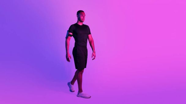 Τρόπος να τέλειο σώμα. Sporty Αφρικής Αμερικανός άνθρωπος με τηλέφωνο Armband Περπάτημα πρόκειται να Γυμναστήριο Γυμναστήριο Γυμναστική σε μωβ φόντο, κοιτάζοντας πέρα με αυτοπεποίθηση έκφραση. Πανόραμα, αντιγραφή χώρου - Φωτογραφία, εικόνα