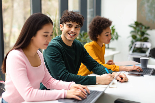 Group of diverse international students sitting at desk in coworking space, focus on smiling guy writing in copybook while preparing for exam or lectures with classmates indoors - Photo, Image