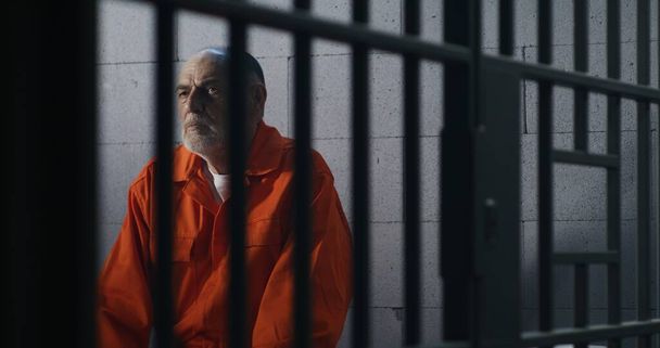 Ältere Kriminelle in orangefarbener Uniform sitzen auf dem Gefängnisbett und denken an die Freiheit. Häftling verbüßt Haftstrafe in Gefängniszelle Schuldiger Häftling in Haftanstalt oder Justizvollzugsanstalt. - Foto, Bild