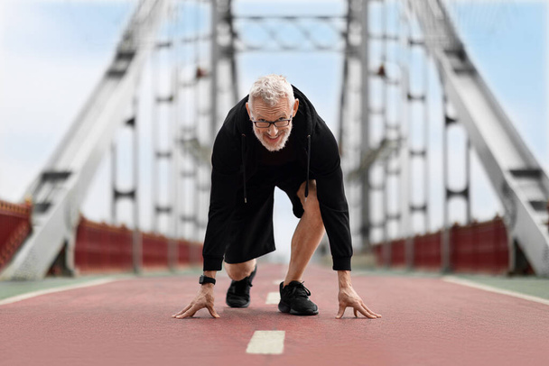 Літній бігун на пенсії стоїть на стартовій робочій позиції на відкритому повітрі. Старший мотивував спортсмена бігати пішохідним мостом, готуючись до марафону. Активний спосіб життя після виходу на пенсію - Фото, зображення
