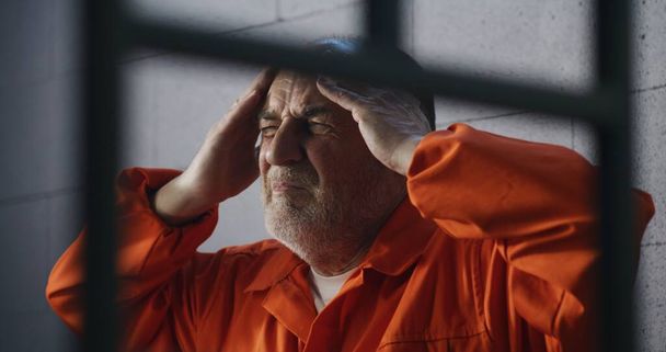 Senior criminal in orange uniform sits on prison bed and dreams about freedom. Prisoner serves imprisonment term in jail cell. Depressed inmate in detention center or correctional facility. - Photo, Image