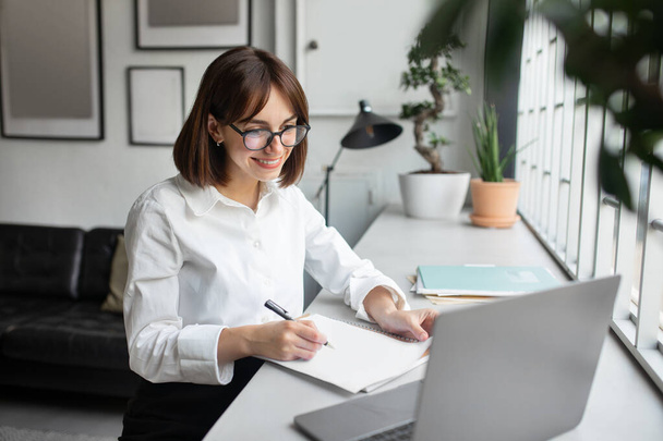 Mujer de negocios sonriente usando el ordenador portátil y tomando notas en el lugar de trabajo en la oficina moderna, señora viendo webinar de negocios y escribiendo algo para copybook, sentado en el escritorio - Foto, imagen