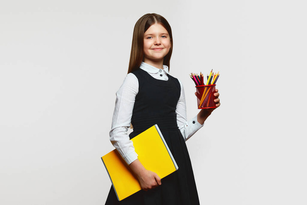 Little pupil girl in school uniform holding notebook and colorful pencils, looking at camera while standing against white background - Фото, изображение