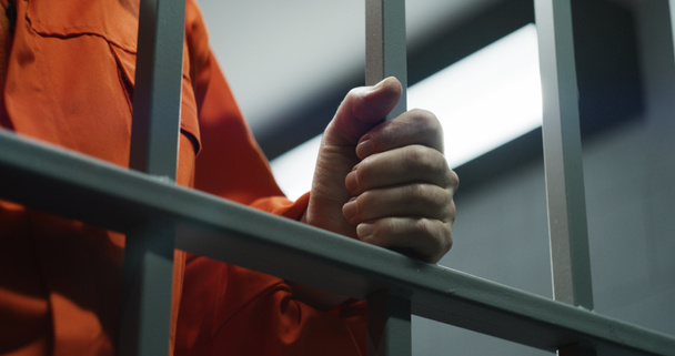 Großaufnahme eines Gefangenen in orangefarbener Uniform mit Metallstangen, der in der Gefängniszelle steht. Schuldiger Verbrecher oder Mörder verbüßt eine Gefängnisstrafe für ein Verbrechen. Häftling im Gefängnis oder in der Haftanstalt. Justizsystem. - Foto, Bild