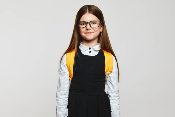 Good looking nerdy pupil kid wearing eyeglasses, school uniform and yellow backpack staring at camera against white background. Back to school concept - 写真・画像