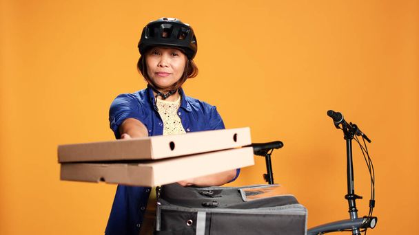 Courier mulher isolado sobre fundo estúdio laranja trazendo pizza para o cliente. Bicicleta piloto BIPOC entregando ordem ao cliente, levando caixas de fast food fora da mochila térmica - Foto, Imagem