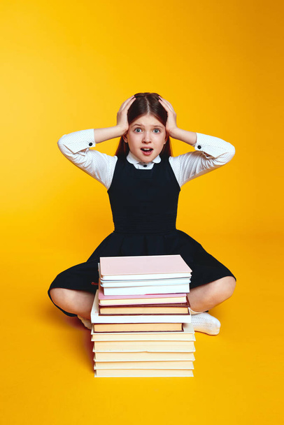 Вертикальное фото изумленной девочки в школьной форме, держащей руки рядом с головой, сидя рядом со стопкой книг, изолированных на желтом фоне - Фото, изображение