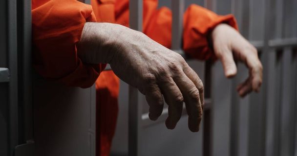 Hände in Großaufnahme eines älteren Häftlings in orangefarbener Uniform, der Metallstangen in der Gefängniszelle hält. Verbrecher verbüßt Freiheitsstrafe für Verbrechen. Häftling im Gefängnis oder in einer Justizvollzugsanstalt. Justizsystem. - Foto, Bild