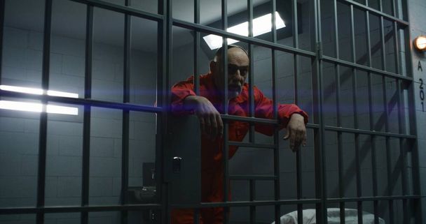 Elderly prisoner in orange uniform leans hands on metal bars. Criminal serves term of imprisonment in prison cell. Gloomy inmate stands behind bars in jail, detention center or correctional facility. - Photo, Image