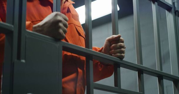 Близько в'язня в помаранчевій формі тримає металеві бруски, що стоять у в'язниці. Винний злочинець або вбивця відбуває термін позбавлення волі за злочин. Ув'язнений у в'язниці або в СІЗО. Система правосуддя. - Фото, зображення