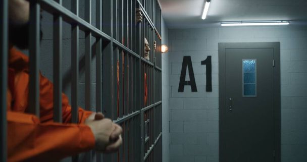 オレンジの制服を着た高齢の囚人は、金属の棒にもたれて指を伸ばします。刑事は刑務所の独房で犯罪の刑期を務める。被収容者は刑務所や拘置所のバーの後ろに立っています. - 写真・画像