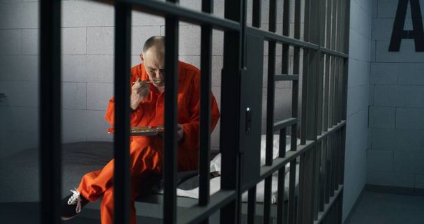 Älterer Häftling, Häftling in orangefarbener Uniform, isst in der Gefängniszelle sitzend Essen. Verbrecher verbüßt Freiheitsstrafe für Verbrechen. Gefängnis, Internierungslager oder Justizvollzugsanstalt. Schießen durch Metallstangen. - Foto, Bild