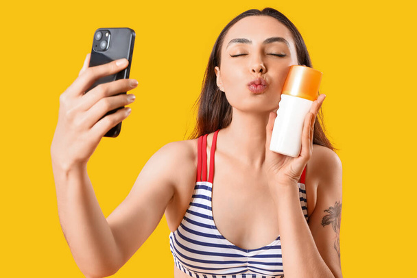 Νεαρή γυναίκα με αντηλιακή κρέμα που παίρνει selfie σε κίτρινο φόντο - Φωτογραφία, εικόνα
