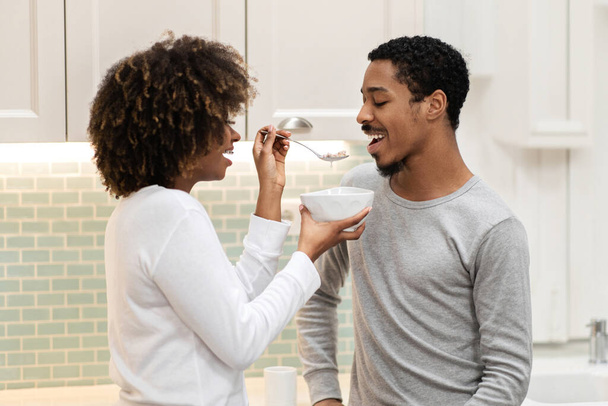 Loving young black woman with bushy hair feeding her handsome boyfriend, wife giving her husband oatmeal. Happy beautiful african american couple have breakfast together at kitchen - Fotografie, Obrázek