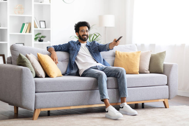 Πορτρέτο του Happy Young Indian Guy βλέποντας τηλεόραση στο σπίτι, χαμογελώντας Millennial Ανατολή άνθρωπος κρατώντας τηλεχειριστήριο και την αλλαγή καναλιών, ενώ χαλαρώνοντας στον καναπέ στο σαλόνι, αντίγραφο χώρου - Φωτογραφία, εικόνα