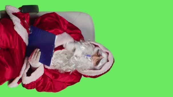 Pystysuora video Edessä näkymä joulupukki mies tekee muistiinpanoja leikepöydälle, kun hän pukeutuu vuodenaikojen punainen puku studiossa. Pyhimys Nickiä esittävä henkilö, joka kirjoittaa tietoja lehtiin, juhlatalvi - Materiaali, video