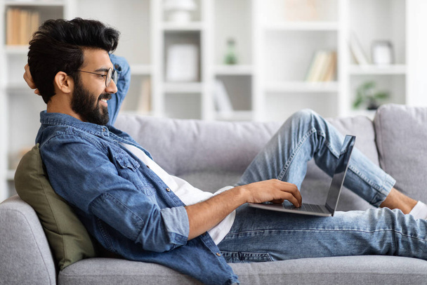 Hálózati koncepció. Indiai srác alkalmi ruhák Laptop használata a nappaliban, jóképű fiatal keleti férfi dolgozik, vagy tanulmány a számítógép, online vásárlás vagy böngészés a szociális hálózatok, Side View - Fotó, kép