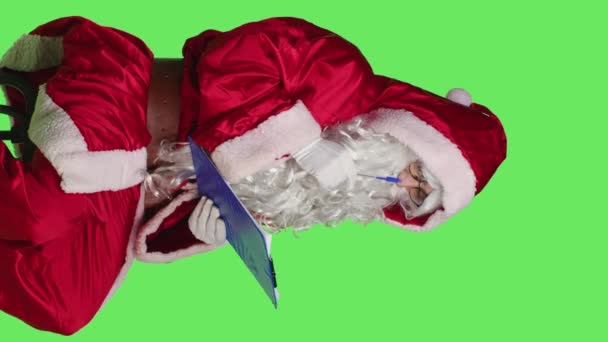 Pionowe wideo Widok z boku człowieka Święty Mikołaj pisać na papierach, siedzi na krześle, podczas gdy on tworzy niegrzeczny i miła lista prezentów. Ojciec Boże Narodzenie przygotowuje się do sezonowej obchodów zimy - Materiał filmowy, wideo