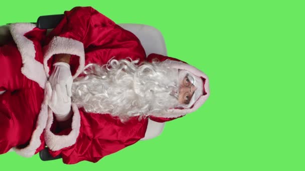 Vertical video Front view of santa claus character on chair against greenscreen backdrop, cheerful person celebrating christmas eve in red costume. Young adult advertising december holiday with iconic - Footage, Video