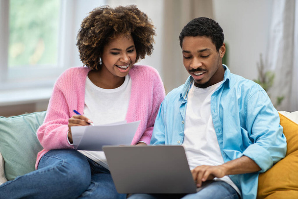 Glücklich fröhlich schöne Millennial schwarzes Paar Mann und Frau auf der Couch sitzen, Papiere füllen und mit PC-Laptop zu Hause. Hypothek, Hauskredit für junge Familien - Foto, Bild