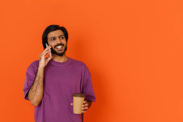 Młody, przystojny, uśmiechnięty Indianin w fioletowej koszulce pijący kawę na wynos podczas przerwy i rozmawiający przez telefon, odizolowany na pomarańczowym tle, patrzący na przestrzeń do kopiowania. - Zdjęcie, obraz