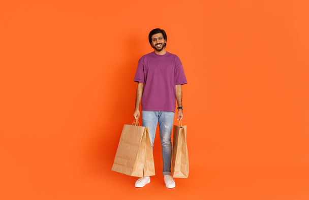 Full length body size view of attractive cheerful eastern guy in casual outfit carrying new things paper bags packages isolated over orange color background, copy space. Shopping, retail concept - Φωτογραφία, εικόνα