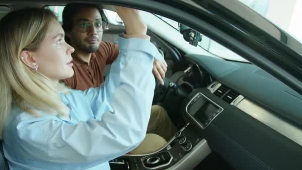 Joven mujer caucásica y su marido de Oriente Medio la elección de un coche nuevo para comprar en concesionario sala de exposición sentado en uno de los modelos de discutir sus características - Metraje, vídeo