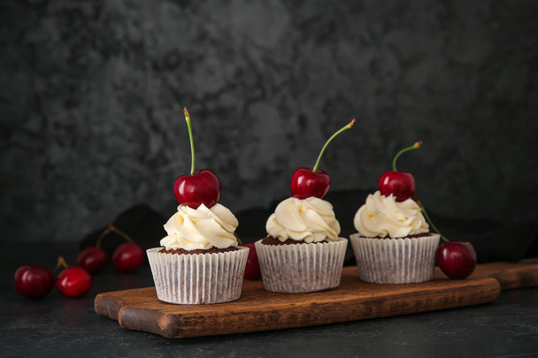 Wooden board with tasty cherry cupcakes on black background - Фото, изображение
