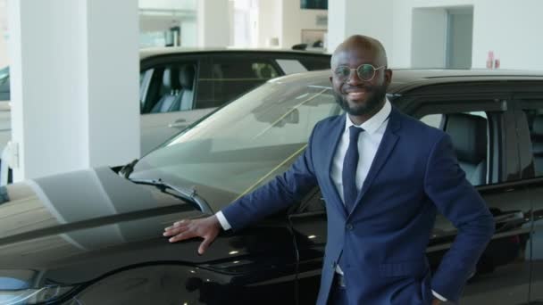 Medium portret van een succesvolle Afro-Amerikaanse man in een elegant pak met een bril tegen een nieuw crossover automodel in de showroom van de dealer glimlachend naar de camera - Video