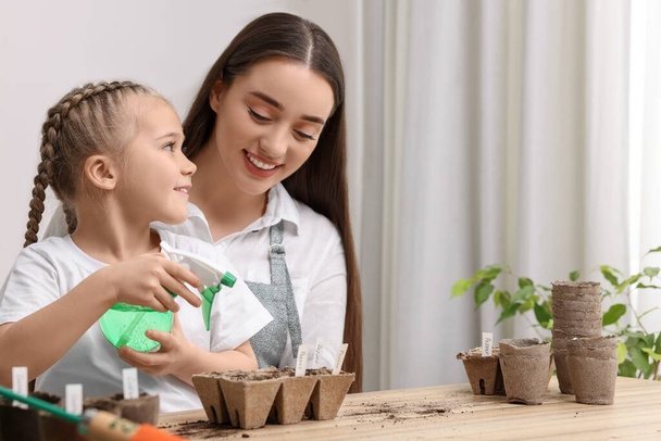 Mother with her daughter spraying water onto vegetable seeds in peat pots at wooden table indoors - Photo, image