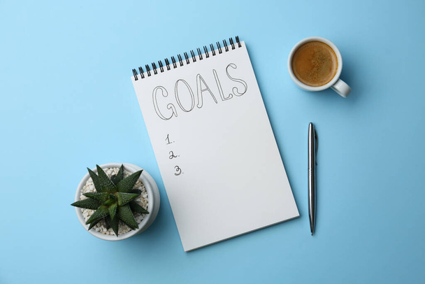 Planning concept. Empty list of goals in notebook, pen, coffee and houseplant on light blue background - Фото, изображение