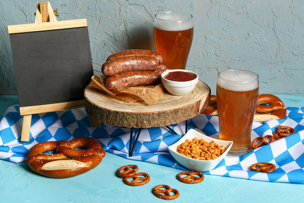 Gläser mit kaltem Bier, leere Tafel und verschiedene Snacks auf blauem Hintergrund. Oktoberfest-Feier - Foto, Bild