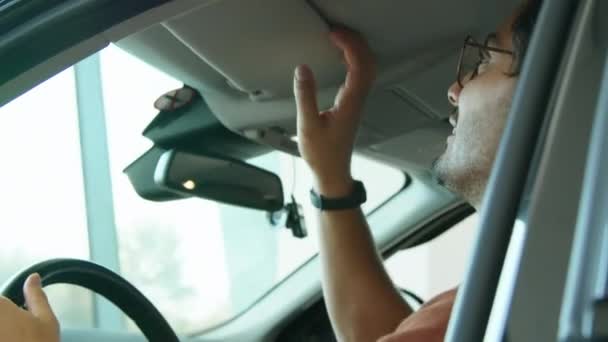 Alegre joven hombre de Oriente Medio sentado en el nuevo modelo de coche en concesionario sala de exposición tocando el volante un visor de sol de comprobación - Metraje, vídeo