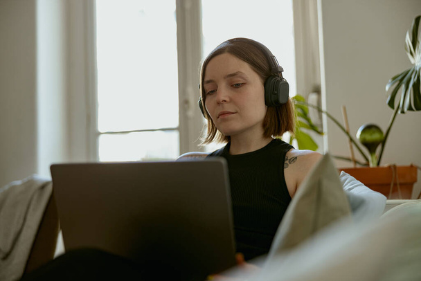 Egy nő fülhallgatóban otthon, kényelmes kanapén fekszik, és laptopot használ. Távolsági munkaterv  - Fotó, kép