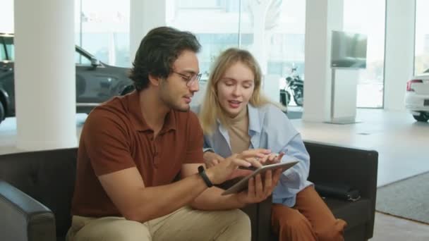 Modern young Middle Eastern man and his Caucasian wife sitting on chairs in dealership showroom holding digital tablet discussing car model features - Footage, Video