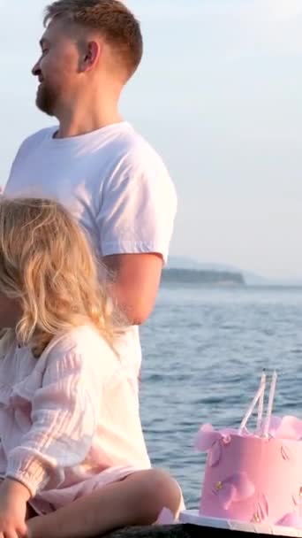 rakkaus ensimmäinen lapsi perheen pikkutyttö blondi kakku 3 vuotta rakkautta vanhempien halaus katsoa toisiaan kivillä lähellä valtameren miesten parta. Vaaleat valkoiset vaatteet Unelmat toteutuvat kaunis perhe - Materiaali, video