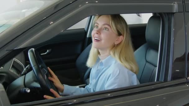 Iloinen nuori valkoihoinen nainen istuu crossover-autossa näyttelytilassa tutkimassa sen sisustusta ja koskettamassa kaikkea siellä - Materiaali, video