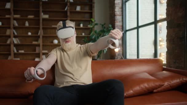 Hipster τύπος φορώντας VR ή γυαλιά εικονικής πραγματικότητας ακουστικά κάθεται σε δερμάτινο καναπέ και παίζει βιντεοπαιχνίδι στο σαλόνι στο σπίτι. Νεαρός γενειοφόρος άνδρας σε casual πουκάμισο με τεχνολογία και καινοτόμο παιχνίδι - Πλάνα, βίντεο