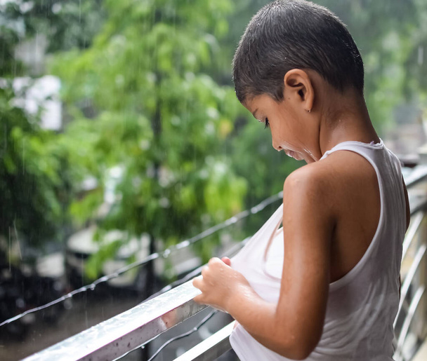 家のバルコニーで夏の雨の中で遊んでいる小さな子供、モンスーン雨の季節の間に雨の滴で遊んでいるインドの賢い男の子、雨の中で遊んでいる子供 - 写真・画像