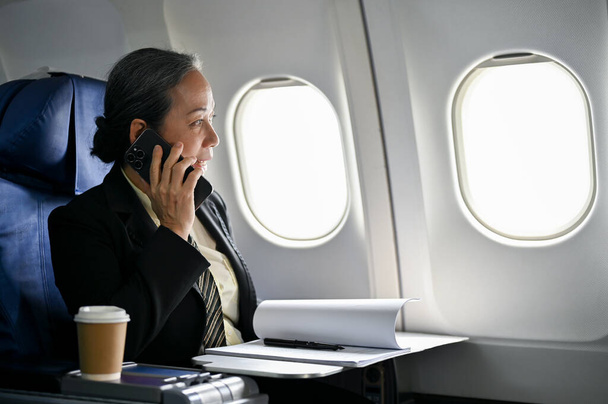 A professional and successful senior Asian businesswoman or female CEO in a formal business suit is on the phone with her business partner during the flight. business trip, airline, journey - 写真・画像