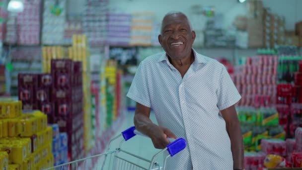 Comprador afroamericano sonriendo a la cámara de pie dentro del supermercado con el carrito de compras. Un consumidor negro brasileño en la tienda de comestibles - Metraje, vídeo